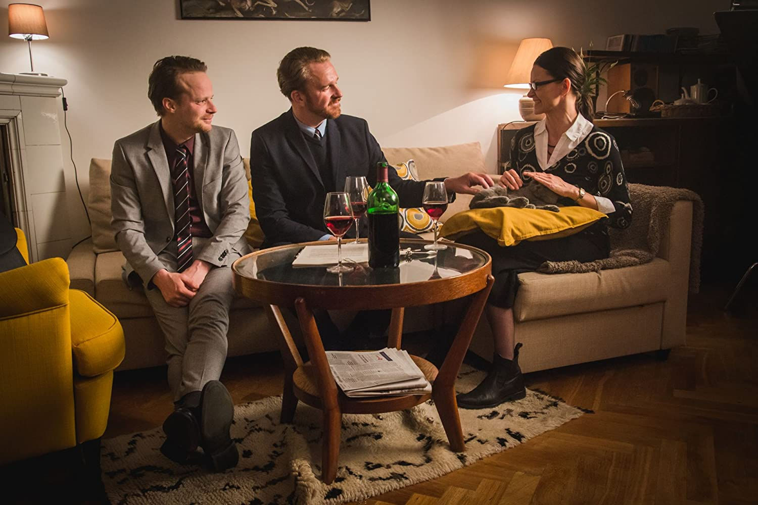 Two men and a woman sitting on a couch, sharing a bottle of wine in a living room.